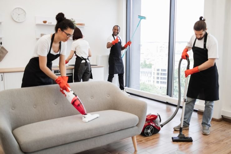 Focus on positive African American worker cleaning panoramic window glass with mop, looking at camera, showing thumb up in modern apartment near multiethnic co-workers.